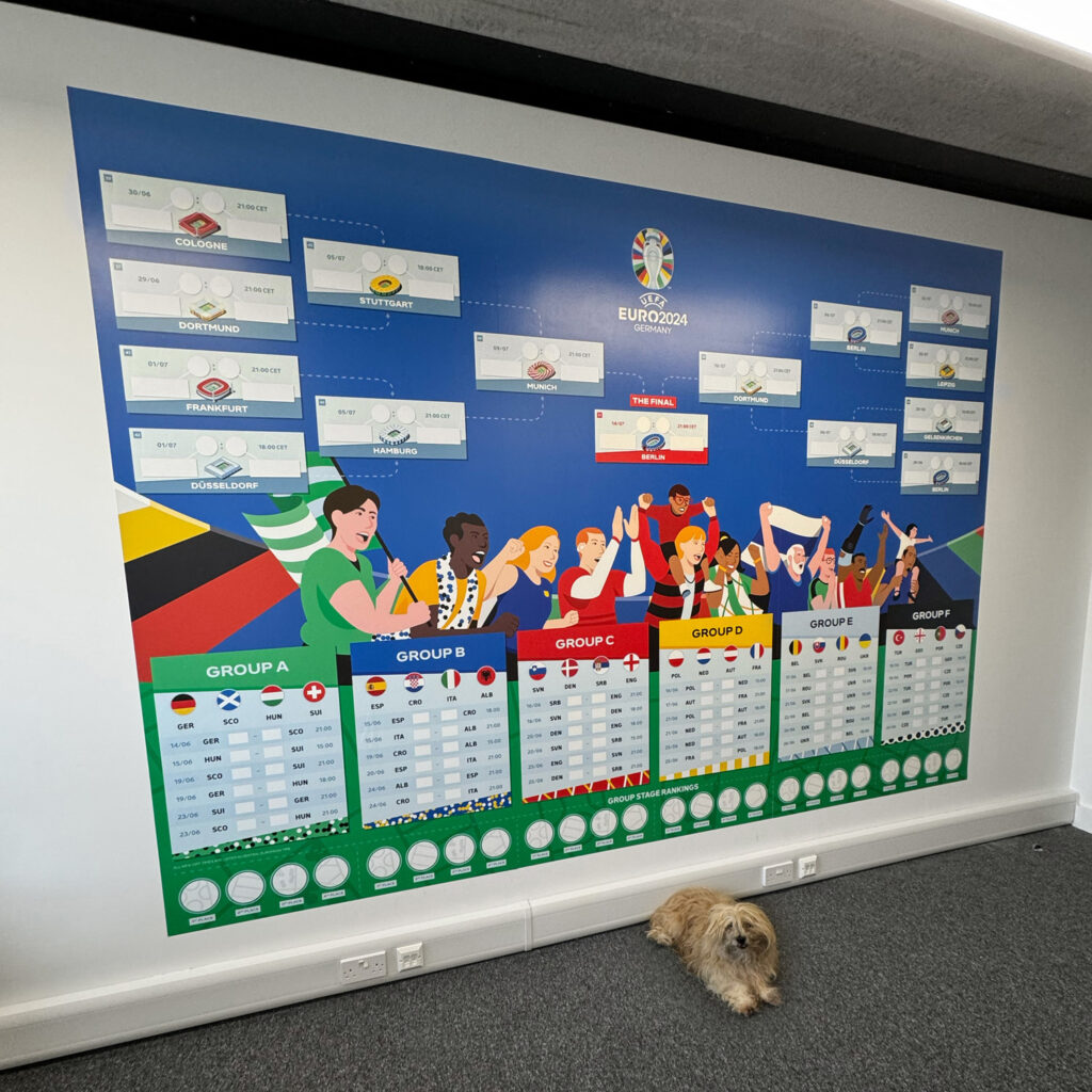 A large Euro 2024 tournament wall chart is displayed on a white wall. The chart shows the tournament bracket, group stages, and match schedules, with colorful graphics of cheering fans representing different countries. At the bottom of the image, a small shaggy dog is lying on the gray carpeted floor in front of the chart.